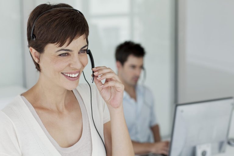 Smiling woman with a headset in an office, with a man working on a computer nearby.