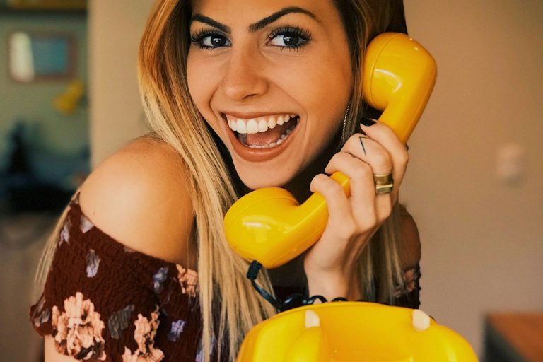 Smiling woman holding a yellow telephone receiver, wearing a floral off-shoulder top.