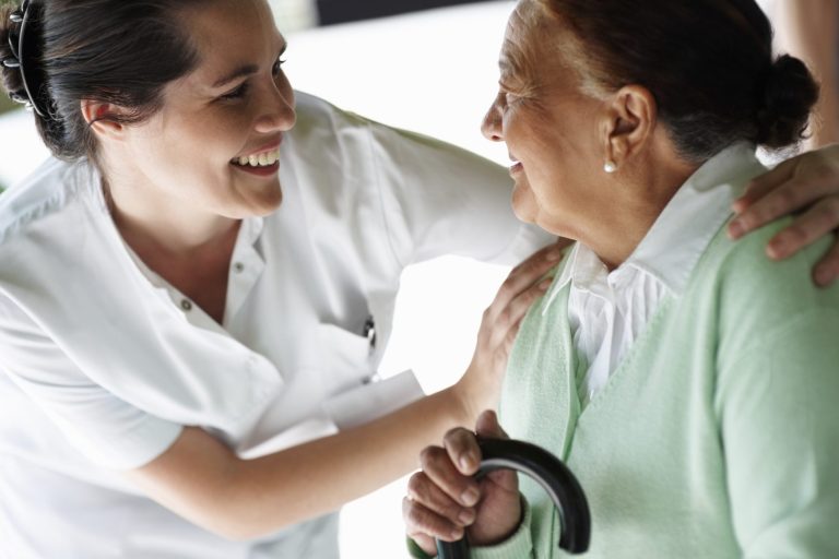 A smiling caregiver interacts warmly with an elderly woman holding a cane.