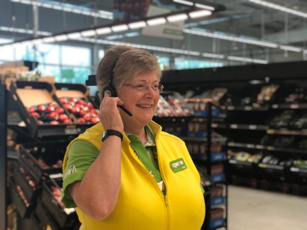 A smiling staff member wearing a yellow vest speaks on a headset in a grocery store.